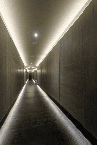 A narrow, modern hallway with wooden walls and a black floor, illuminated by a continuous strip light on the ceiling and recessed spotlights.