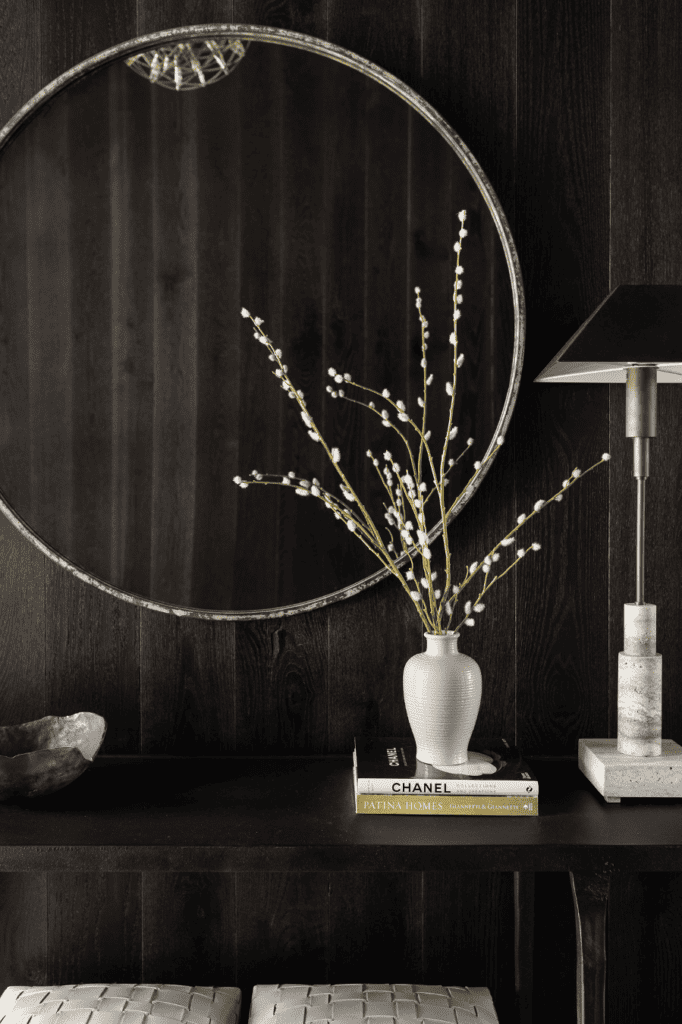 A stylish interior with a circular mirror, white vase with branches, books, a stone bowl, a concrete lamp, and a dark wooden background.