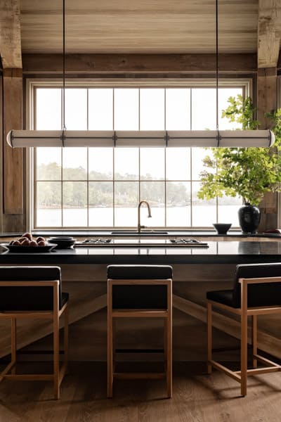 Modern kitchen with wooden cabinetry, a linear suspension light, a gas cooktop, and three chairs aligned with a central island. Large window with outdoor view.
