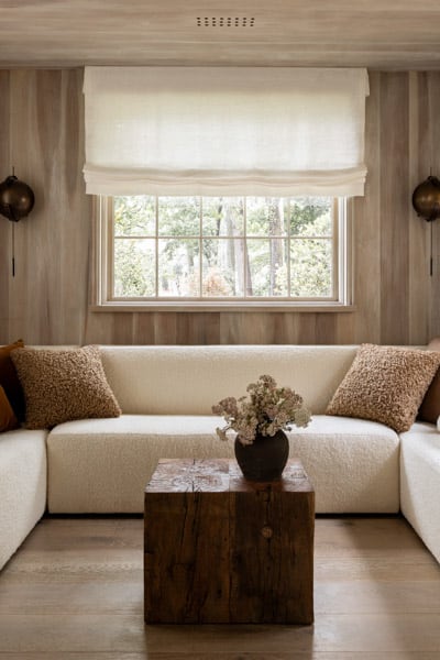 A cozy room featuring a cream corner sofa with plush pillows, a rustic wooden coffee table, flanked by wall sconces, under a large window with a roman shade.