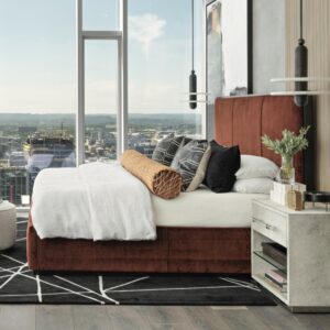 This image shows a modern bedroom with a large window offering a city view, a stylish bed with pillows, and contemporary decor elements. From an article about Textures Nashville featured in Luxe magazine.