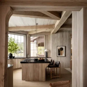 A modern kitchen with wooden exposed beams, light wood cabinets, a central island, and sleek chairs set in a clean, minimalist design. Large windows provide natural light. From an article in Architectural Digest titled 6 Design Collabs We’re Loving Right Now featuring the new Jeffrey Dungan line by Textures Nashville.