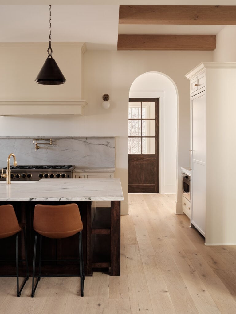 Elegant kitchen with marble countertops, wooden beams, white cabinetry, and a single pendant light. Two stools under the island, hardwood floors throughout.