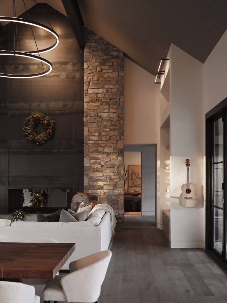 Modern living space with stone wall, concrete panels, and wooden ceiling. Cozy sofa, dining area, decorative wreath, and guitar on white wall visible.