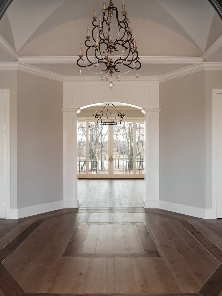 An elegant interior with herringbone wooden flooring leading to an arched doorway, ornate chandeliers above, and a view of a glass-paneled balcony.