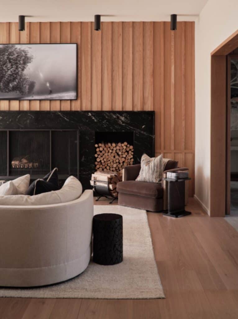 A cozy living room with a circular beige sofa, wooden walls, a black marble fireplace, a monochrome photograph, and stacked firewood.