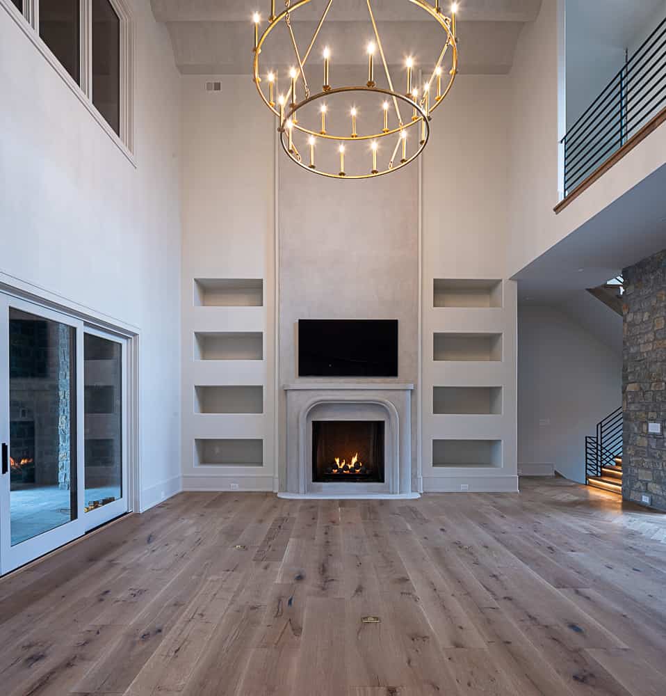 The image shows a modern spacious living room with high ceilings, a large fireplace, built-in shelves, and an elegant circular chandelier.